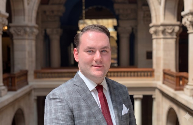 A man in a gray suit and red tie standing in a grand, ornate interior.