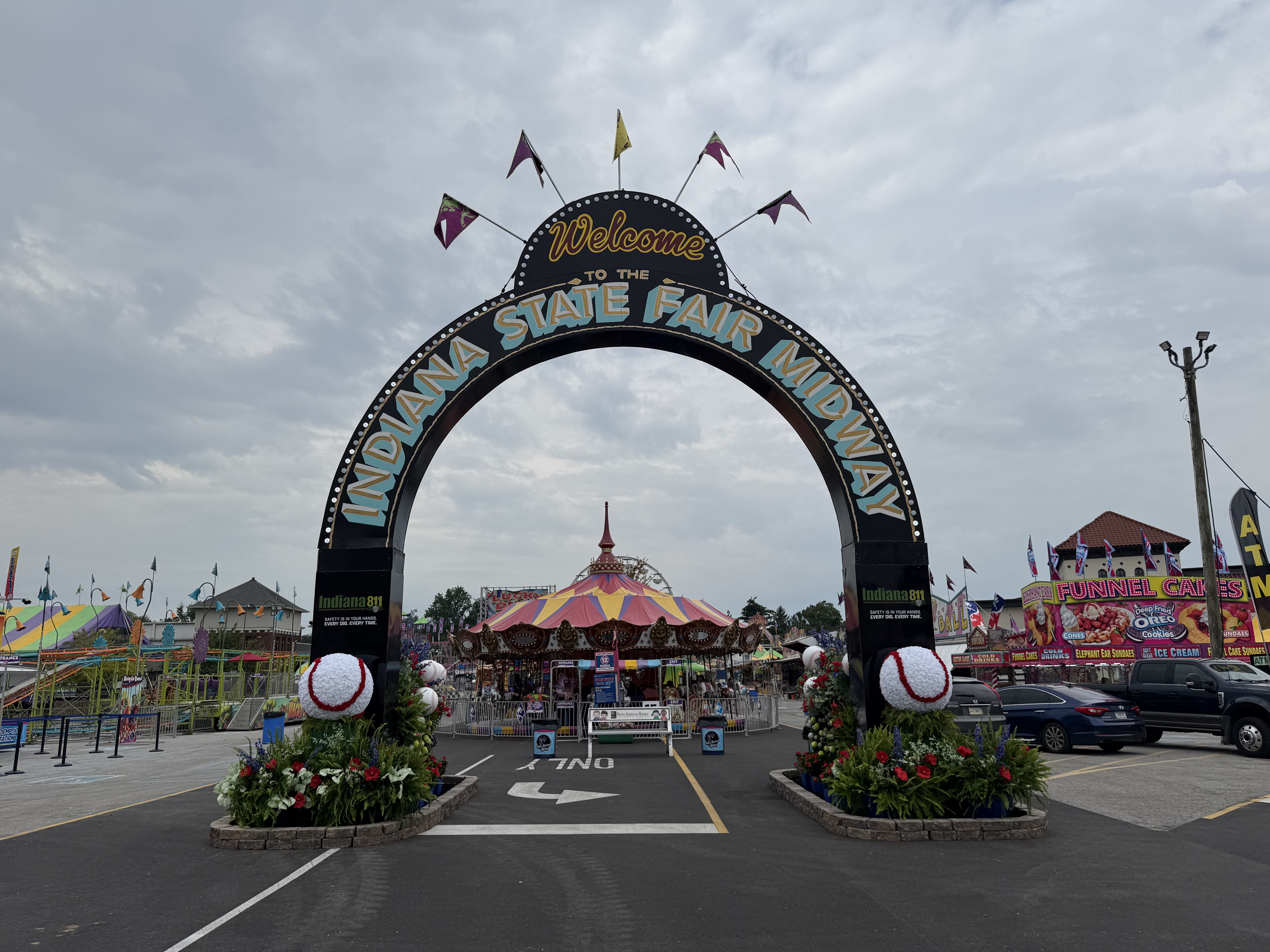 Welcome to the Indiana State Fair Monday arch with a colorful carnival scene behind it, including a large tent-like structure and various rides and attractions.