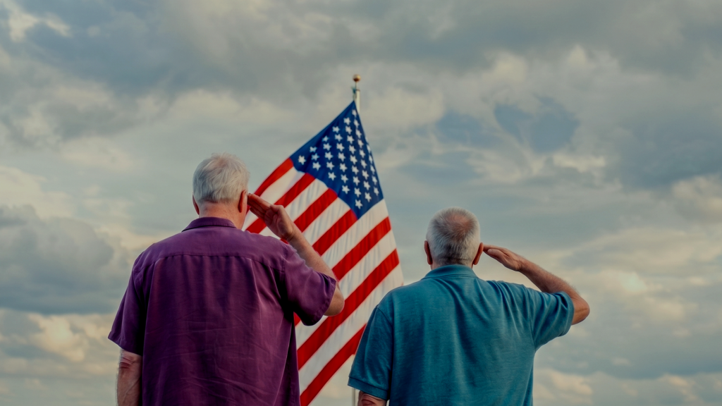 Two older men standing and saluting the American flag against a cloudy sky.