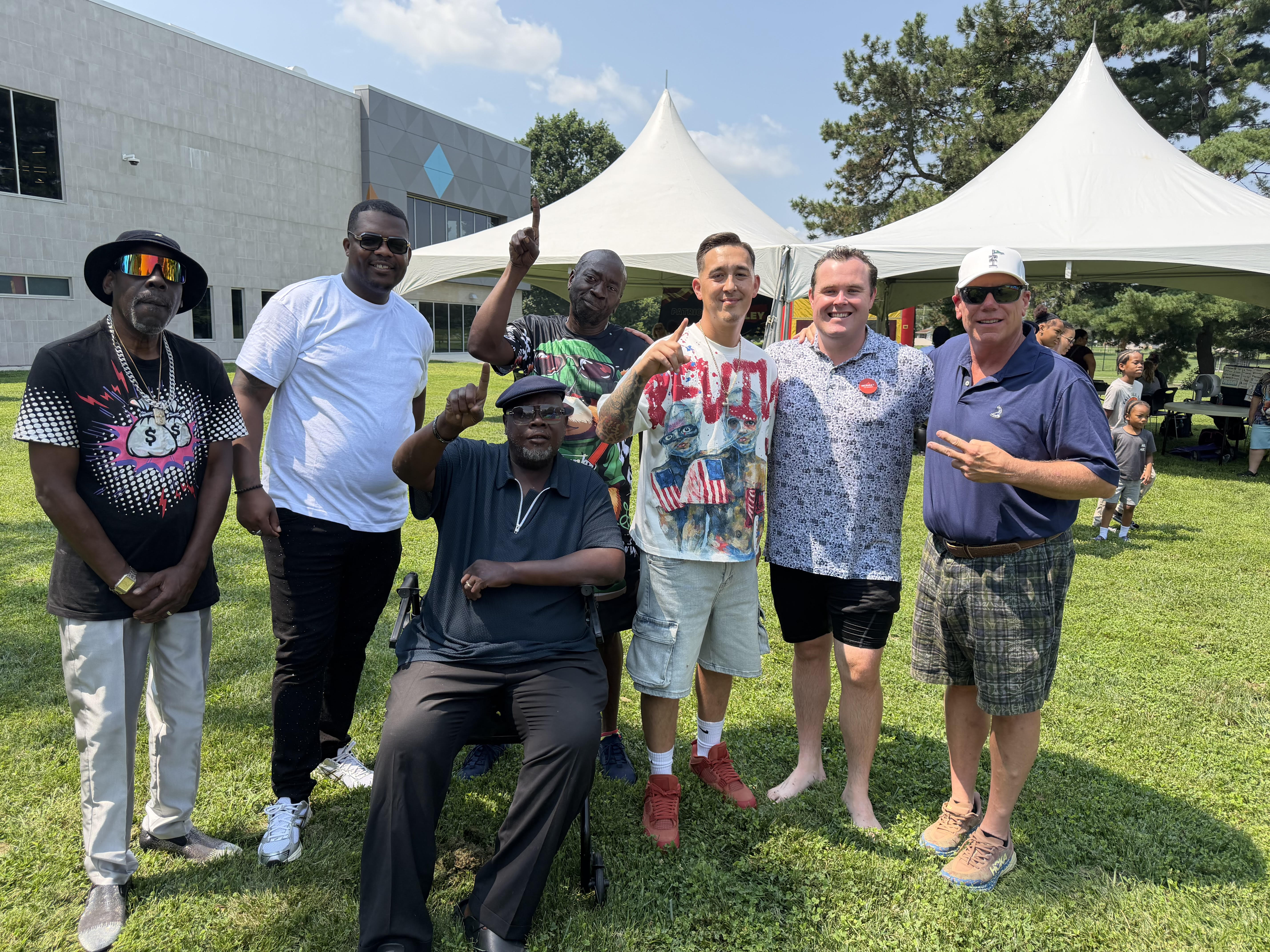 A group of diverse people posing together outdoors, with tents and trees in the background. One person is wearing a shirt with the word "REVOLT" printed on it.