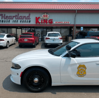 A white police car parked in front of a jewelry store called "King Jewelers" and a retail store called "Heartland".