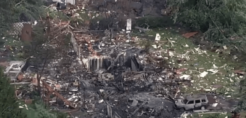 Aerial view of a destroyed residential area with debris, damaged buildings, and a vehicle visible.
