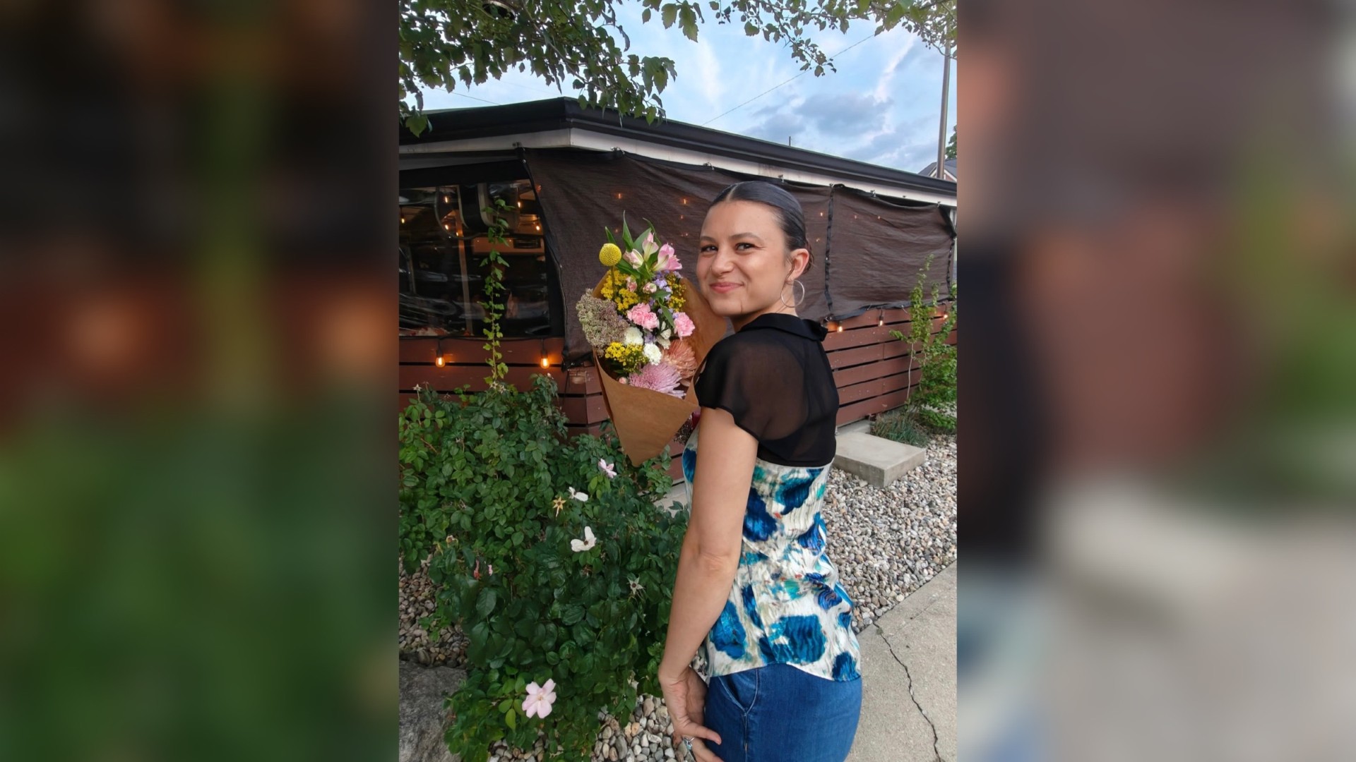A smiling woman holding a bouquet of flowers in front of a wooden structure with string lights.