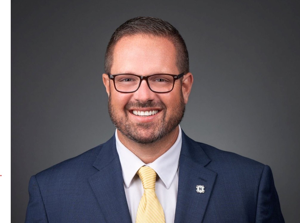 A smiling man with a beard wearing glasses, a navy blue suit, and a yellow tie.