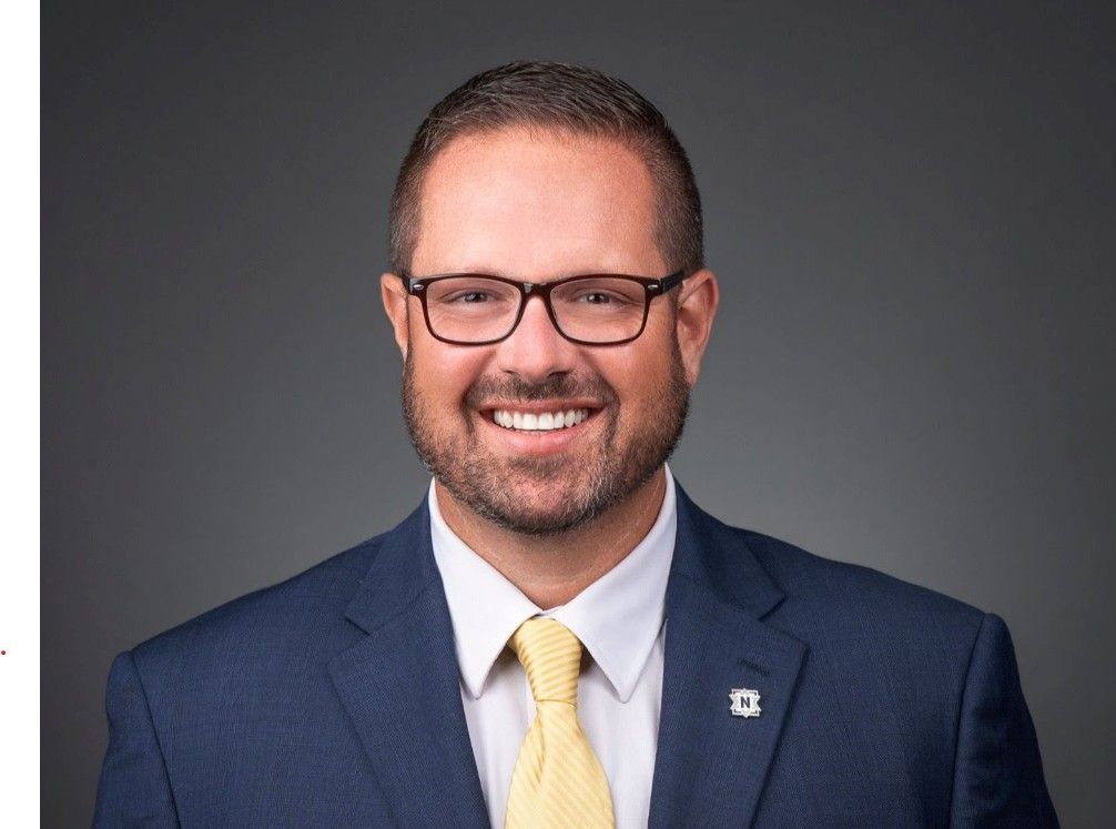 A smiling man with a beard wearing glasses, a navy blue suit, and a yellow tie.