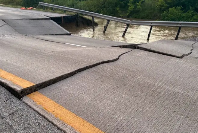 Damaged road with large cracks and gaps, likely caused by flooding or erosion, with a guardrail visible in the background.