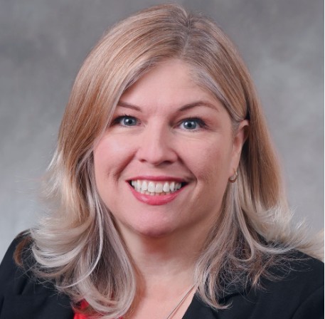 A smiling woman with blonde hair wearing a black blazer against a gray background.