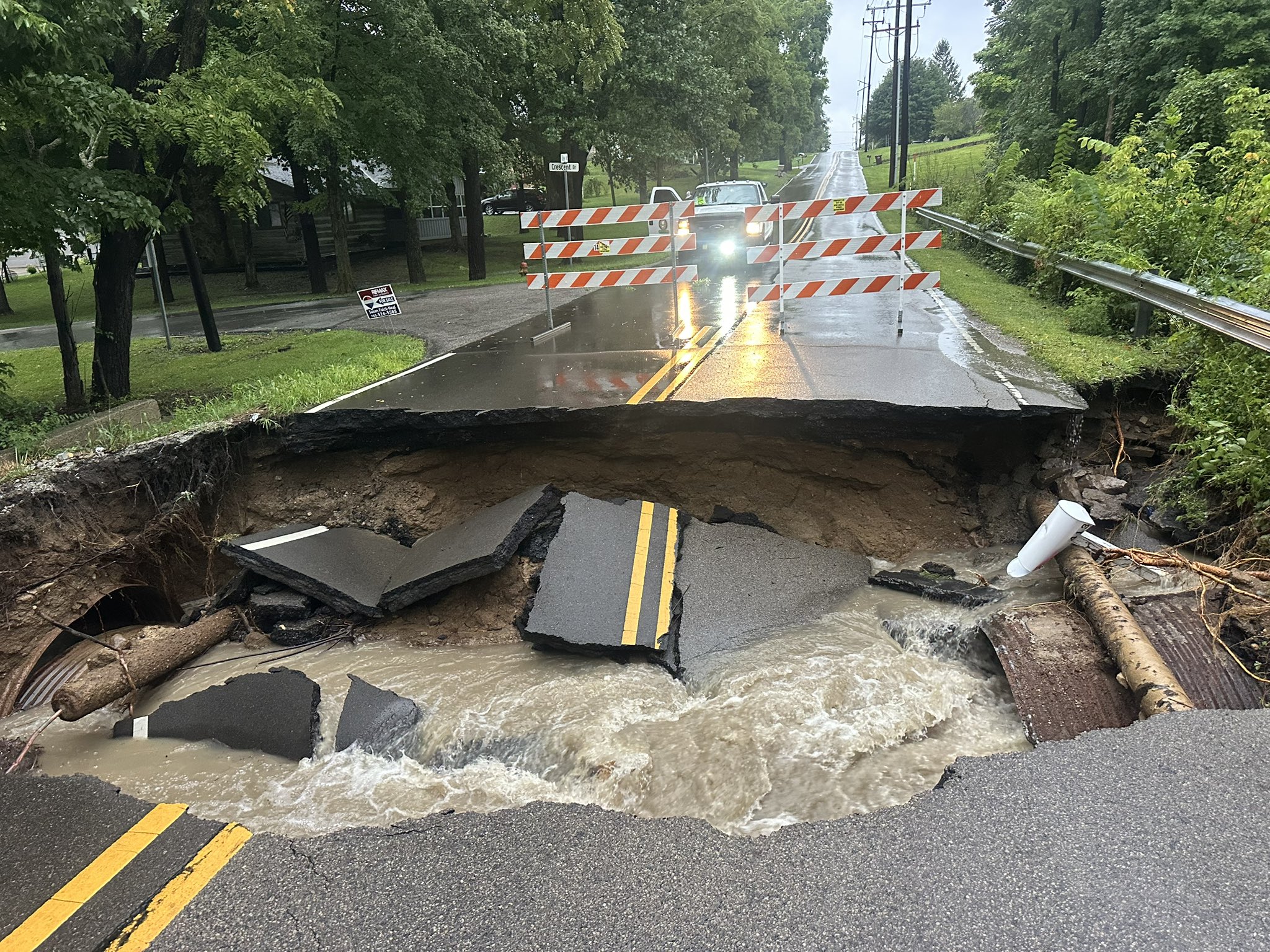 A road has been washed out by heavy rain, with debris and a damaged barrier blocking the way. A utility truck is visible in the distance.