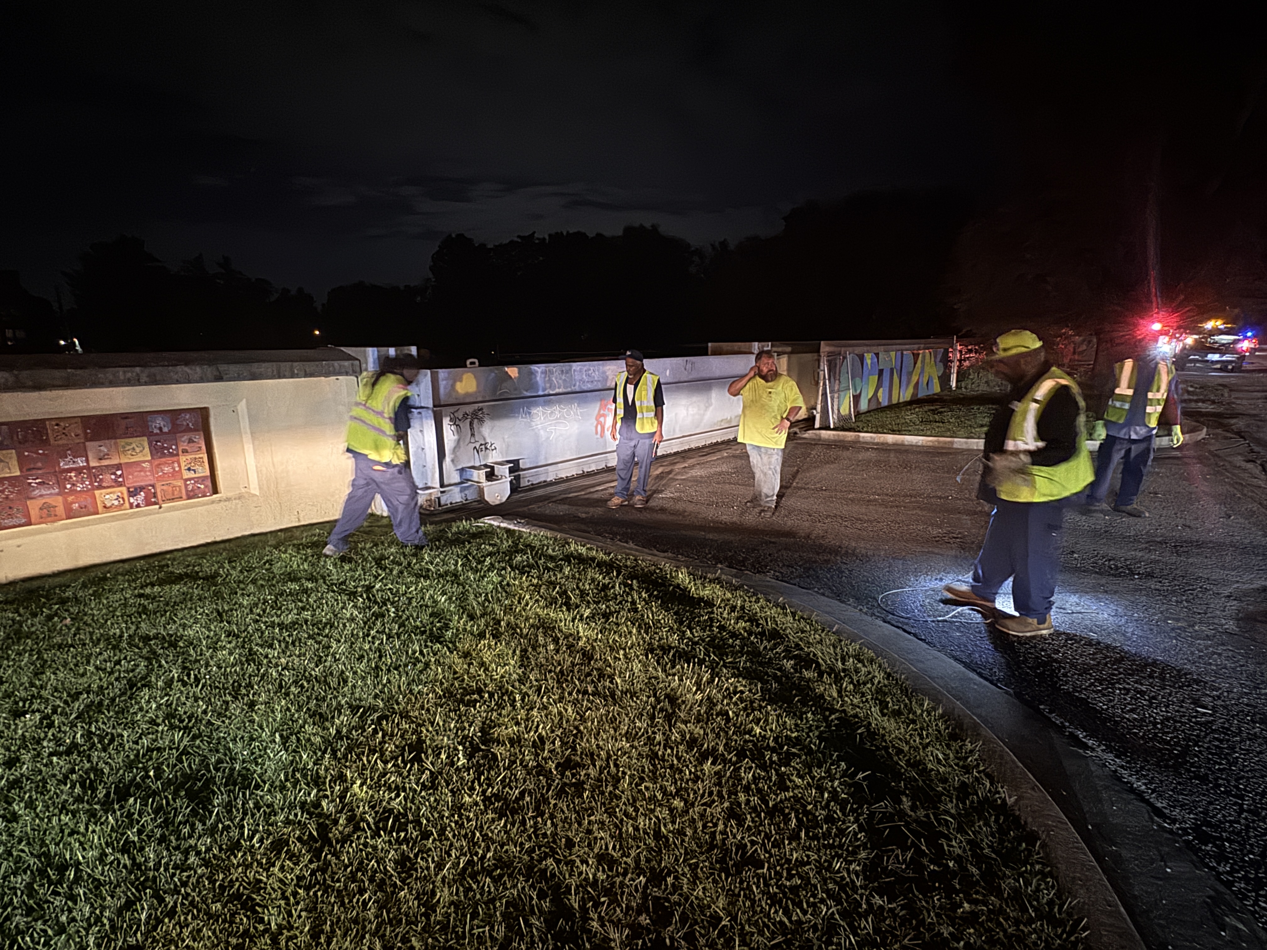 A group of construction workers in high-visibility vests working at a construction site at night, with a large illuminated sign visible in the background.