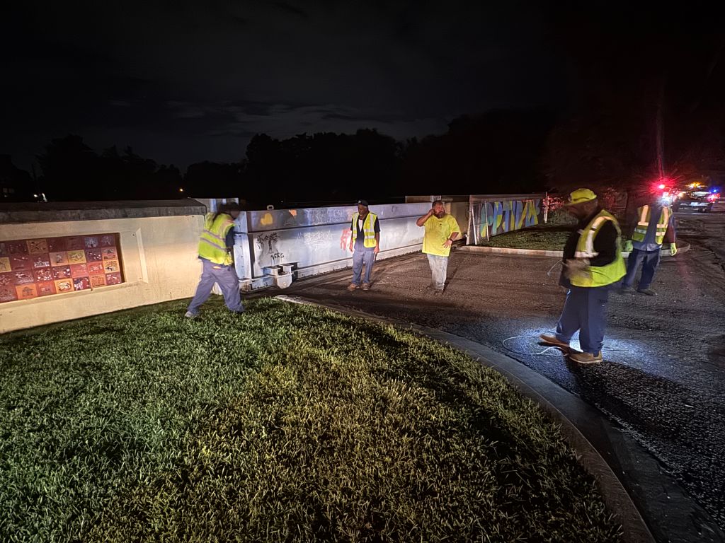 A group of construction workers in high-visibility vests working at a construction site at night, with a large illuminated sign visible in the background.