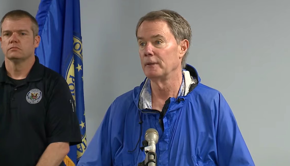 Two men standing in front of a blue flag with a state seal, one wearing a police uniform and the other speaking into a microphone.
