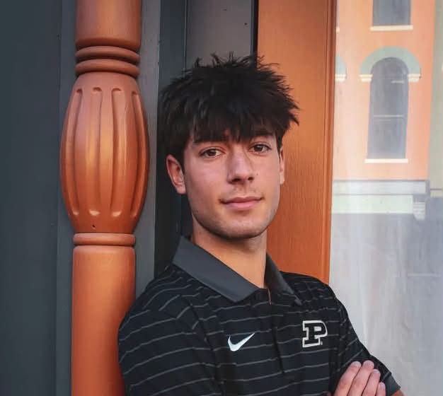 A young man wearing a black and white striped Nike polo shirt stands with his arms crossed in front of a wooden door.