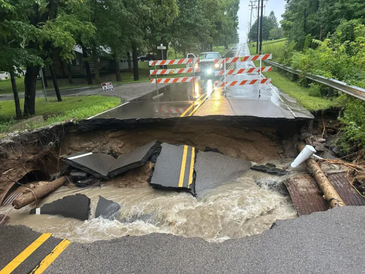 A road has been washed away by floodwaters, with debris and damage visible. Barricades and warning signs are set up around the damaged area.