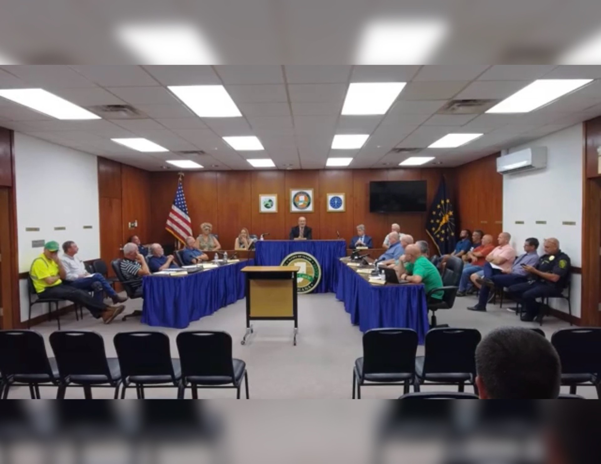 A government meeting room with officials seated around a table, an American flag, and a podium with a seal displayed.