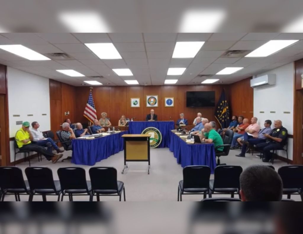 A government meeting room with officials seated around a table, an American flag, and a podium with a seal displayed.