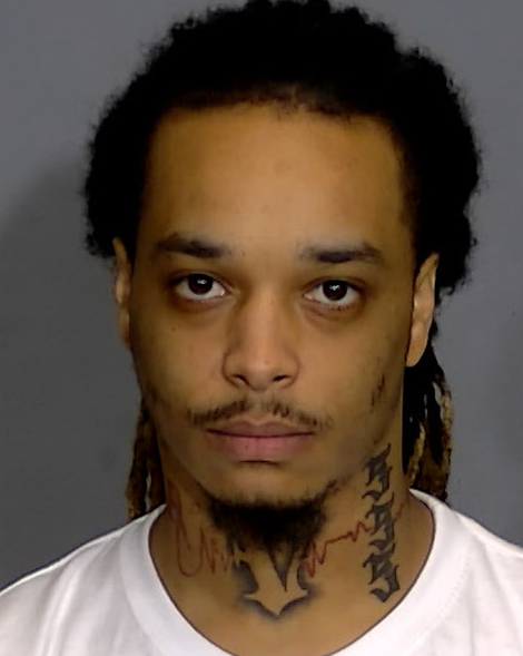 A close-up portrait of a man with dreadlocks and facial tattoos, looking directly at the camera with a serious expression.