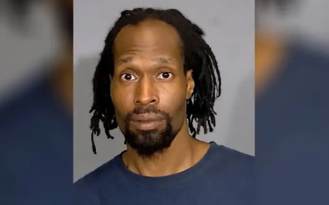 Headshot of a man with dreadlocks and a beard, wearing a navy blue shirt, looking directly at the camera with a serious expression.