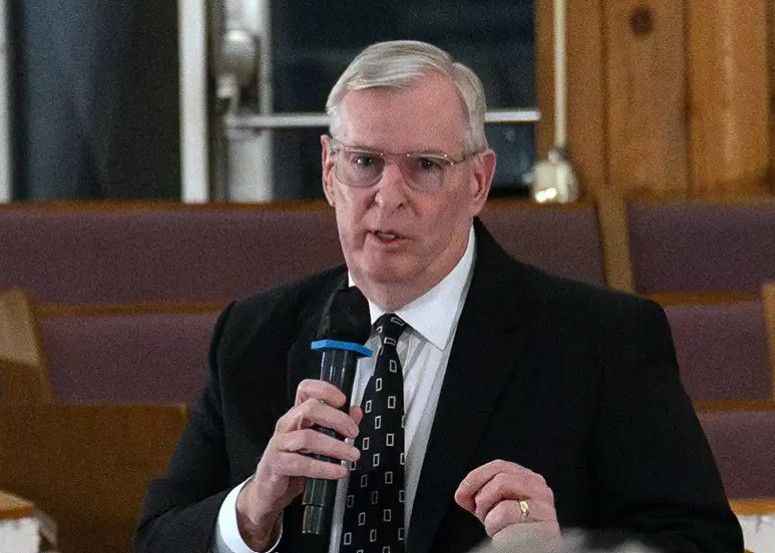 An older man in a suit speaking into a microphone on a stage.