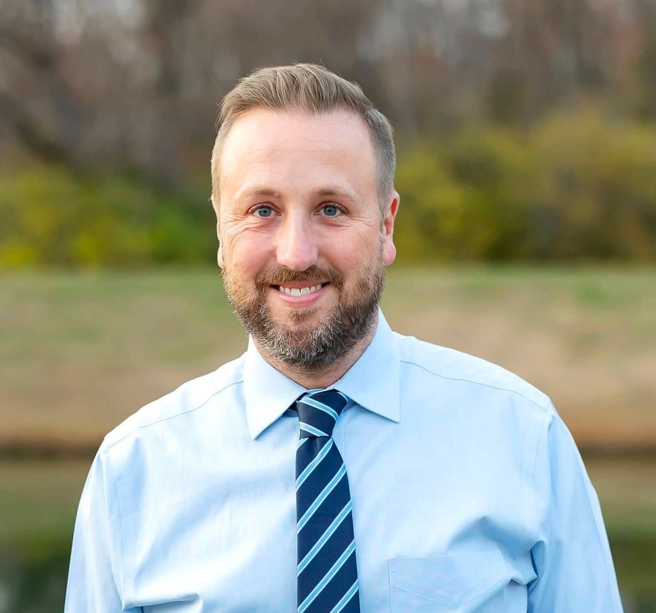 A smiling man with a beard wearing a light blue shirt and navy blue tie, standing outdoors in a natural setting.