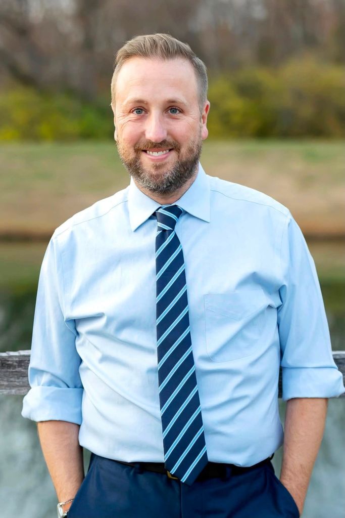 A smiling man with a beard wearing a light blue shirt and navy blue tie, standing outdoors in a natural setting.