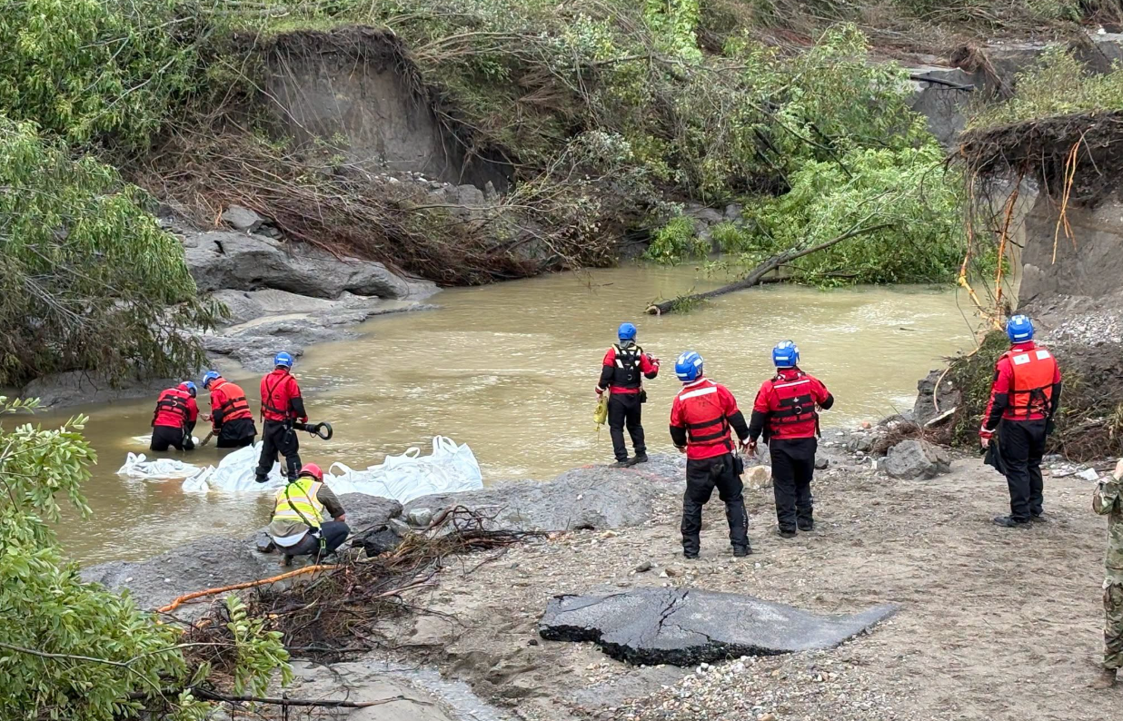 A group of rescue workers in red and blue uniforms standing in a river surrounded by debris and vegetation.