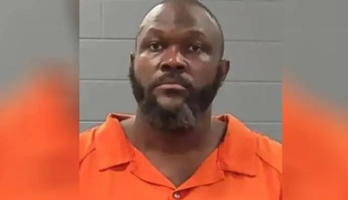 A man with a beard wearing an orange prison uniform looks directly at the camera.