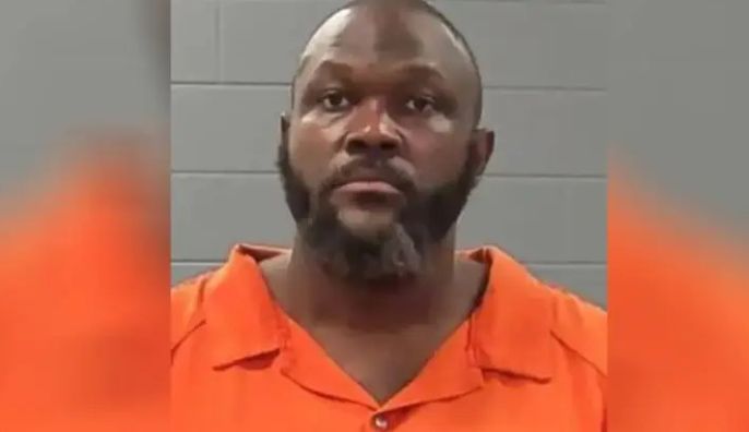 A man with a beard wearing an orange prison uniform looks directly at the camera.