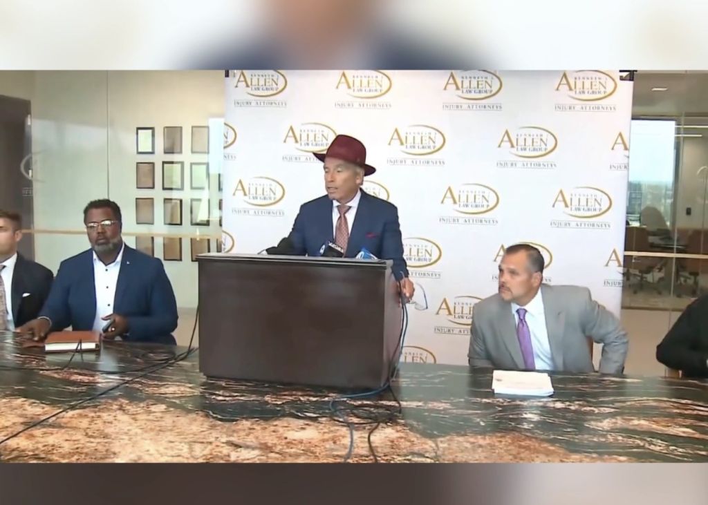 A man in a burgundy hat speaking at a podium with "Allen & Associates" branding behind him, surrounded by other men in suits.