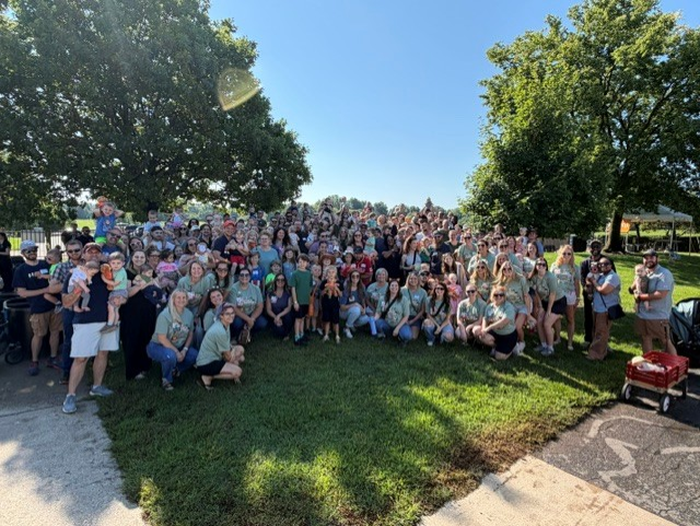A large group of people gathered outdoors on a sunny day, wearing matching green shirts and posing for a group photo.