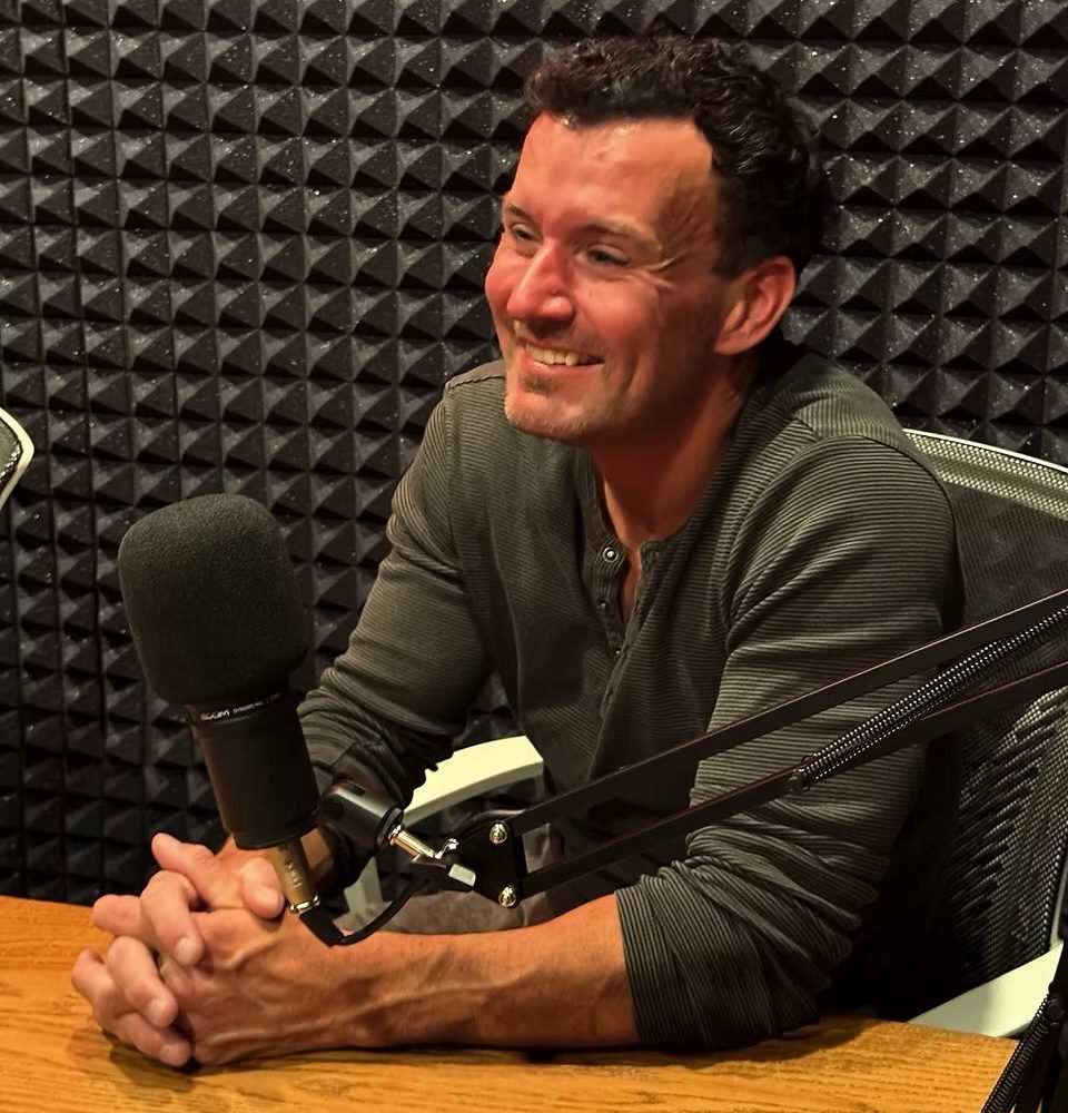 A smiling man wearing a gray shirt sits at a microphone in a recording studio with a patterned wall behind him.