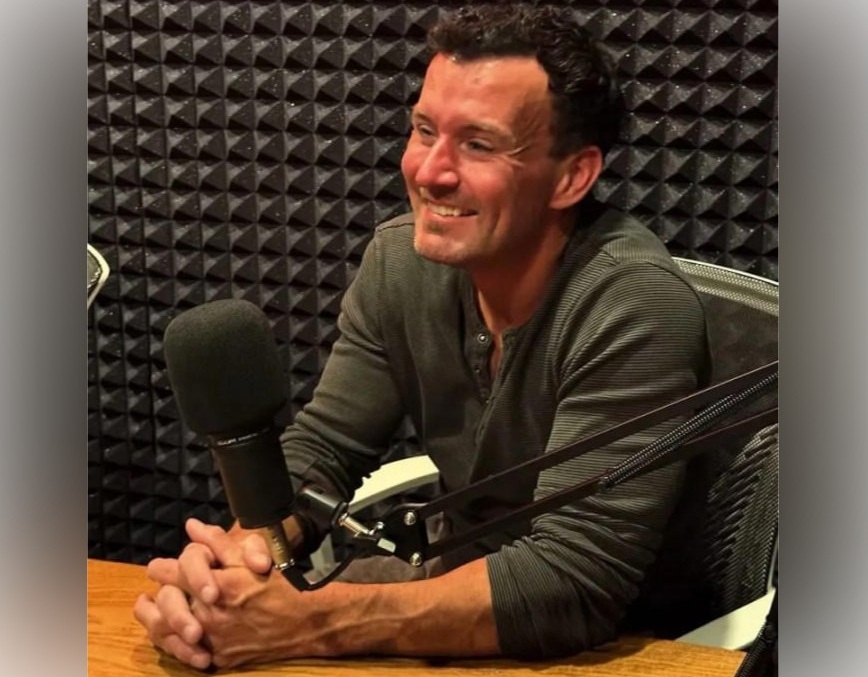 A smiling man sitting at a microphone in a recording studio.