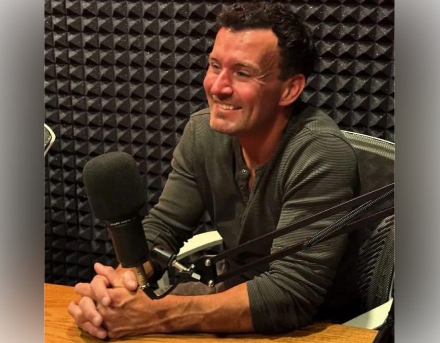 A smiling man sitting at a microphone in a recording studio.