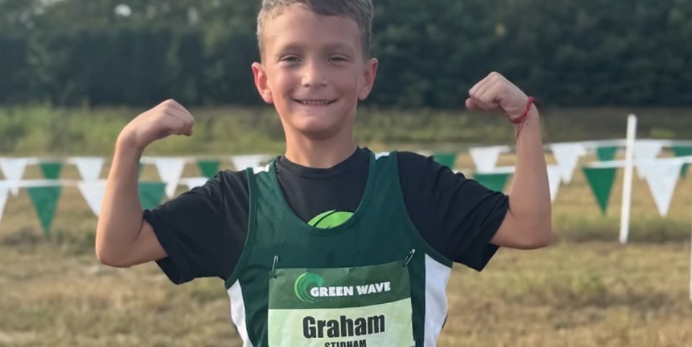 A smiling young boy wearing a green "Green Wave" jersey and flexing his muscles while standing in a field with green flags in the background.