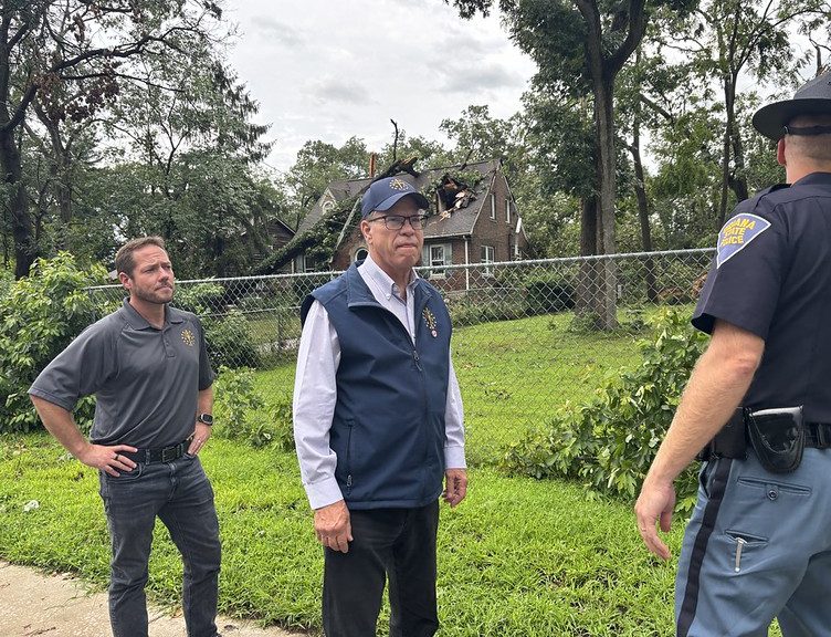 Three men, one wearing a police uniform, standing in a grassy area near a damaged building.