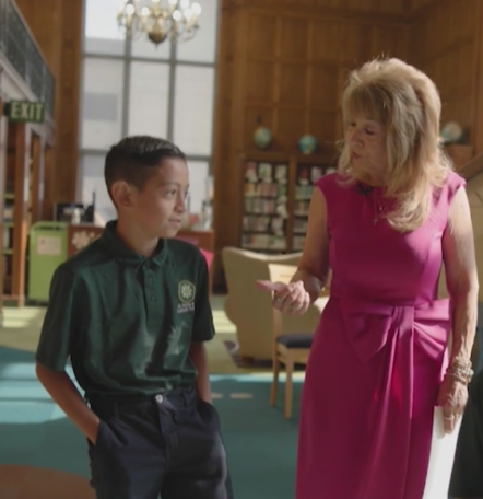 A young boy in a green uniform standing and talking to an older woman in a pink dress in what appears to be a school or community center setting.