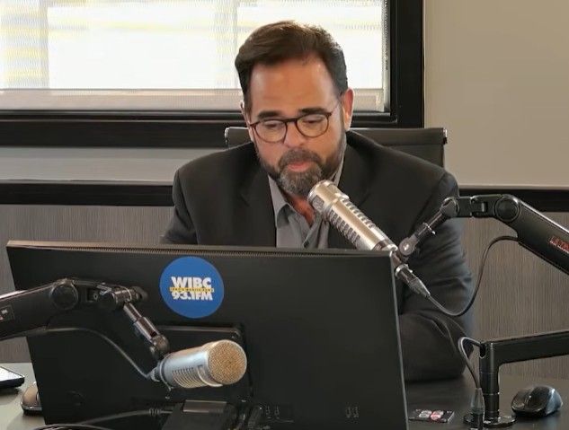 A man with a beard and glasses speaking into a microphone at a radio studio, with a WIBC logo visible on the equipment.