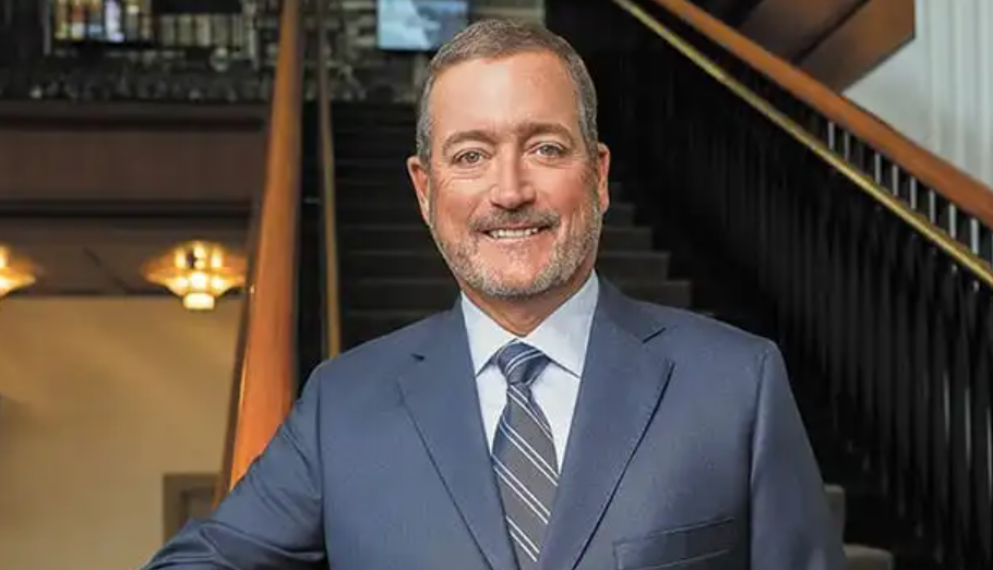 A smiling middle-aged man in a navy blue suit and tie standing in a room with a bar and shelves in the background.