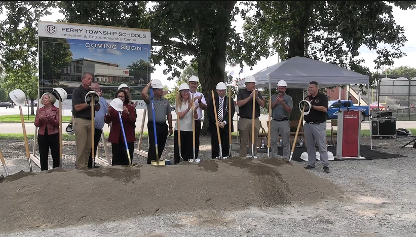 A group of people, including school administrators and construction workers, standing in front of a "Perry Township Schools" sign and a construction site, holding shovels and preparing for a groundbreaking ceremony.