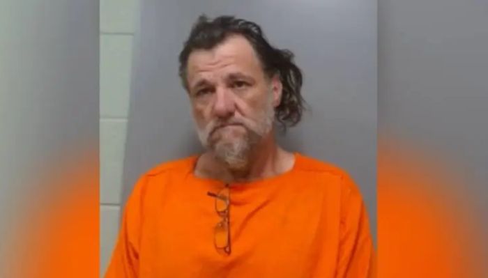 A middle-aged man with a beard wearing an orange prison uniform, standing against a gray wall.