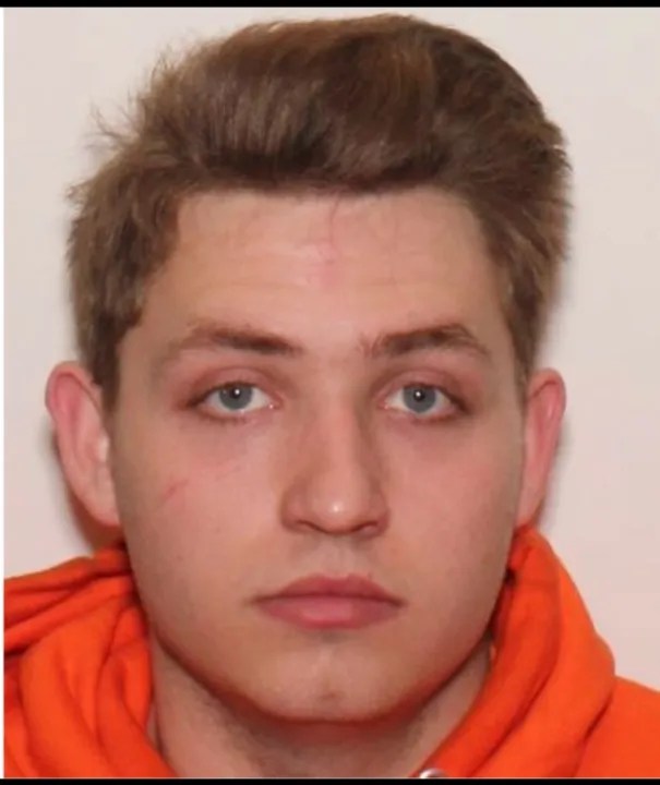 A young man with short brown hair wearing an orange shirt looks directly at the camera with a serious expression.